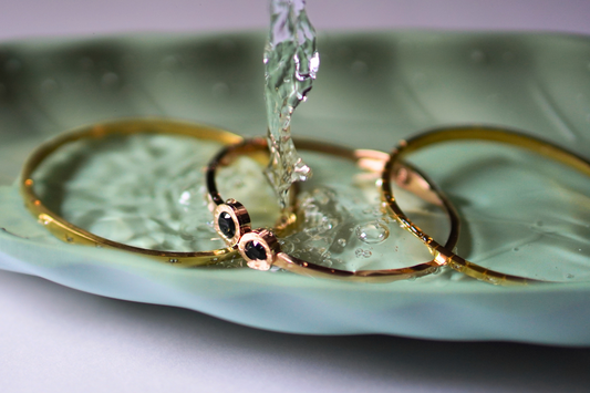 Elegant gold bangles, including one with black gemstone accents, rest on a pastel green ceramic dish as a stream of water pours over them, creating a luxurious and refreshing aesthetic.