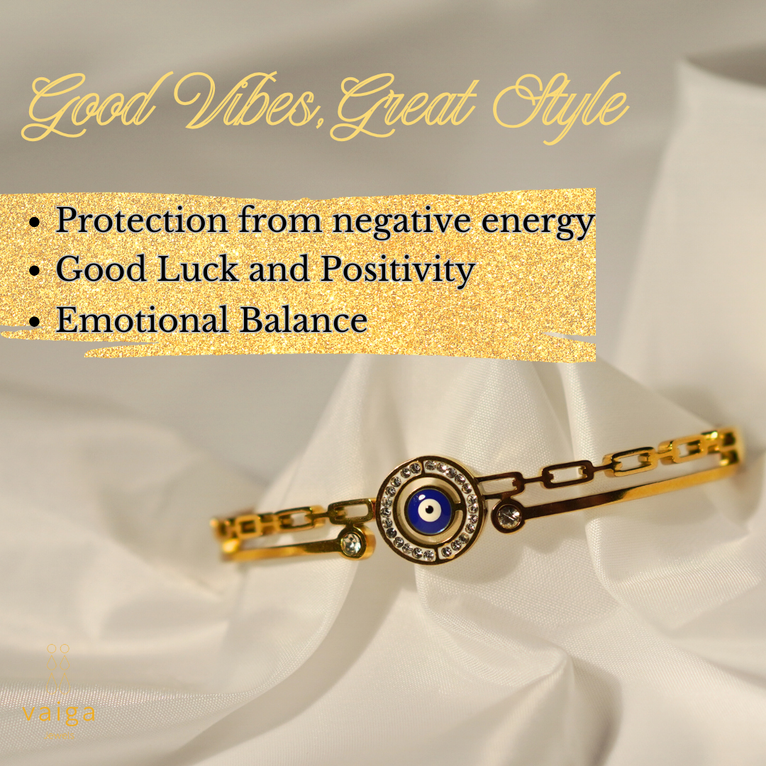Gold-plated stainless steel evil eye bracelet with crystal accents, elegantly displayed on white fabric. The image features text highlighting its benefits: protection from negative energy, good luck, and emotional balance. Vaiga Jewels branding in the corner.
