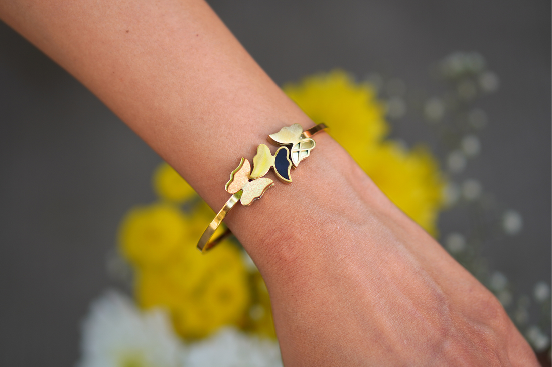 A close-up of a woman's wrist adorned with a gold bangle bracelet featuring delicate butterfly-shaped embellishments with enamel detailing. The background consists of blurred yellow and white flowers, adding a soft and elegant touch to the image.