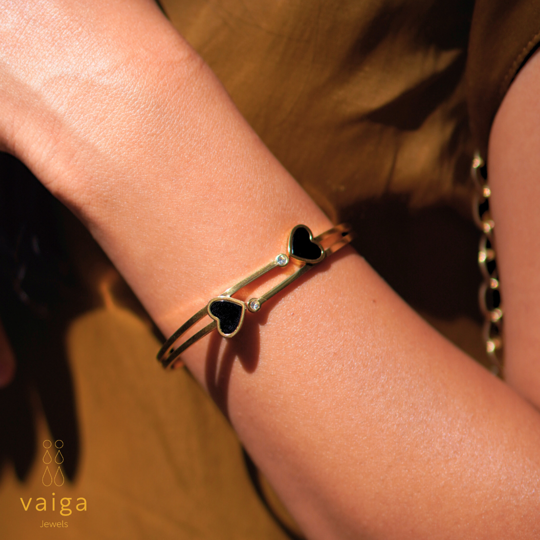 A gold-toned bracelet with black heart-shaped elements and small clear gemstones is elegantly worn on a wrist. The bracelet features a double-band design, adding a modern and stylish touch. The background showcases a warm-toned outfit, complementing the jewelry. The logo "Vaiga Jewels" is visible in the bottom left corner, enhancing the branding of the image.