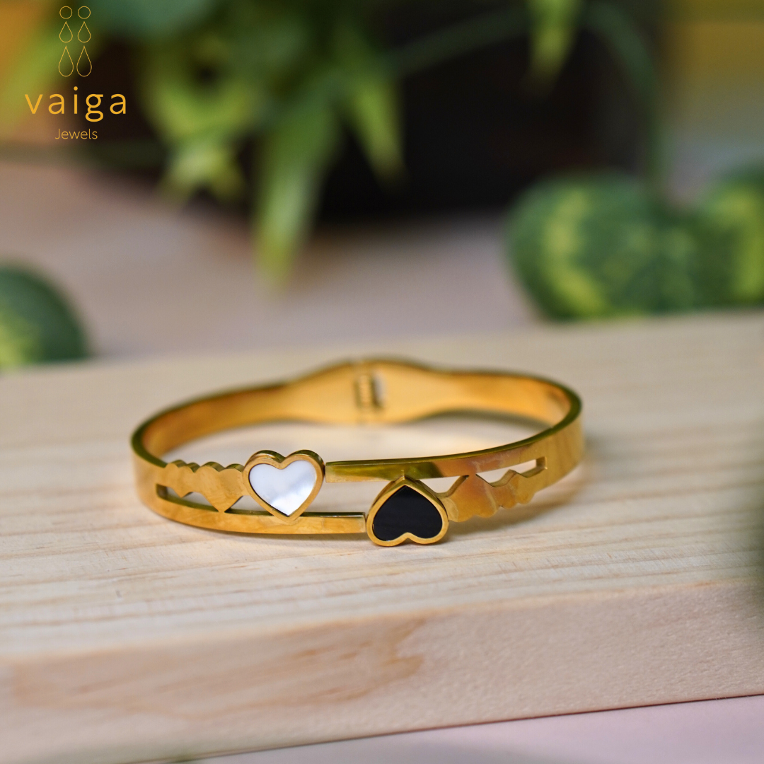  A gold-toned bracelet with an elegant heart-themed design, featuring a white enamel heart and a black enamel heart set within an openwork pattern of butterflies and geometric shapes. The bracelet rests on a wooden surface with a blurred background of green plants, adding a natural and sophisticated touch. The "Vaiga Jewels" logo is placed in the upper left corner.