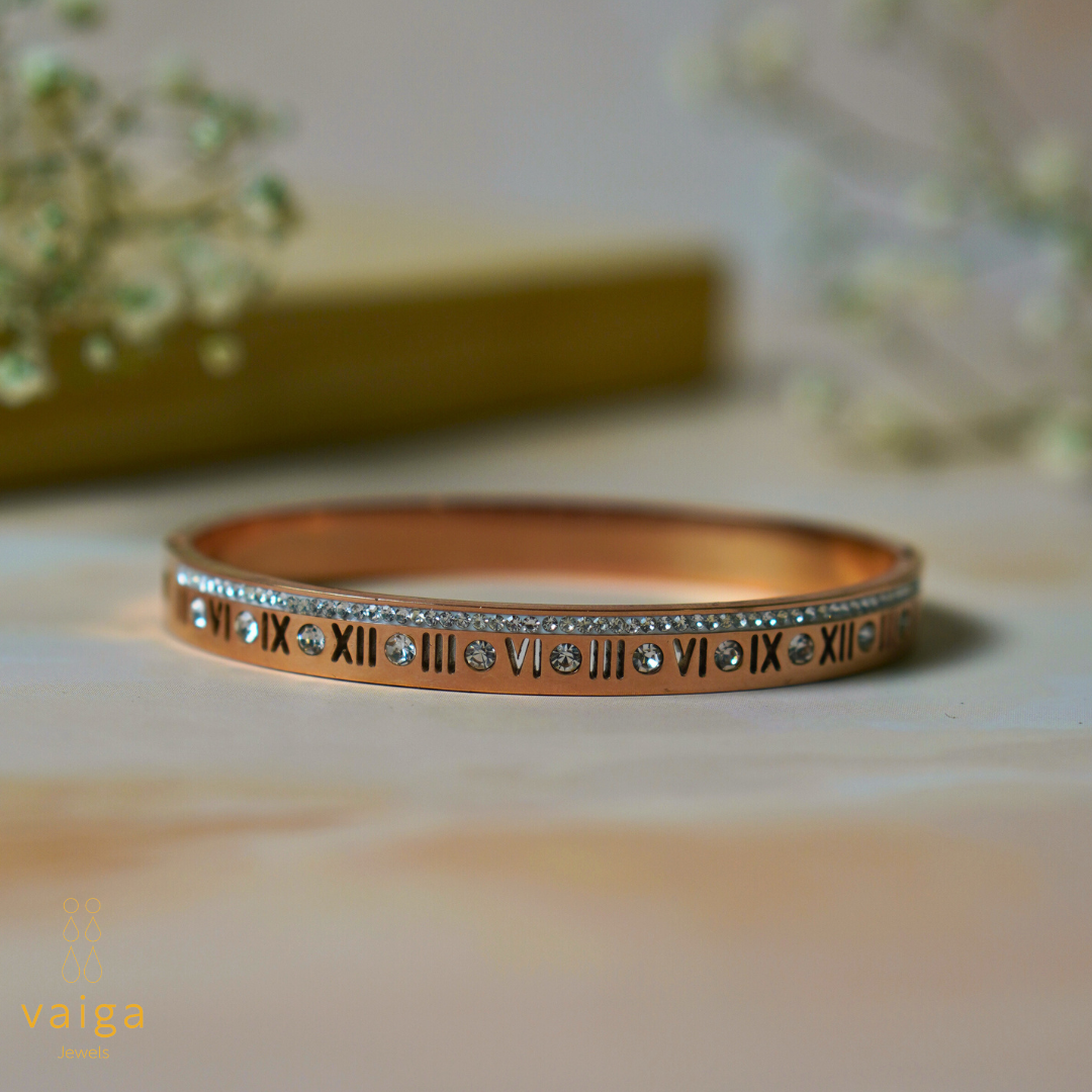 A rose gold-toned bracelet featuring Roman numerals and embedded clear gemstones is elegantly displayed against a soft, neutral backdrop. A row of sparkling stones lines the top edge, adding a touch of luxury. The bracelet's sleek and modern design makes it a timeless piece. The "Vaiga Jewels" logo in the bottom left corner enhances brand recognition.