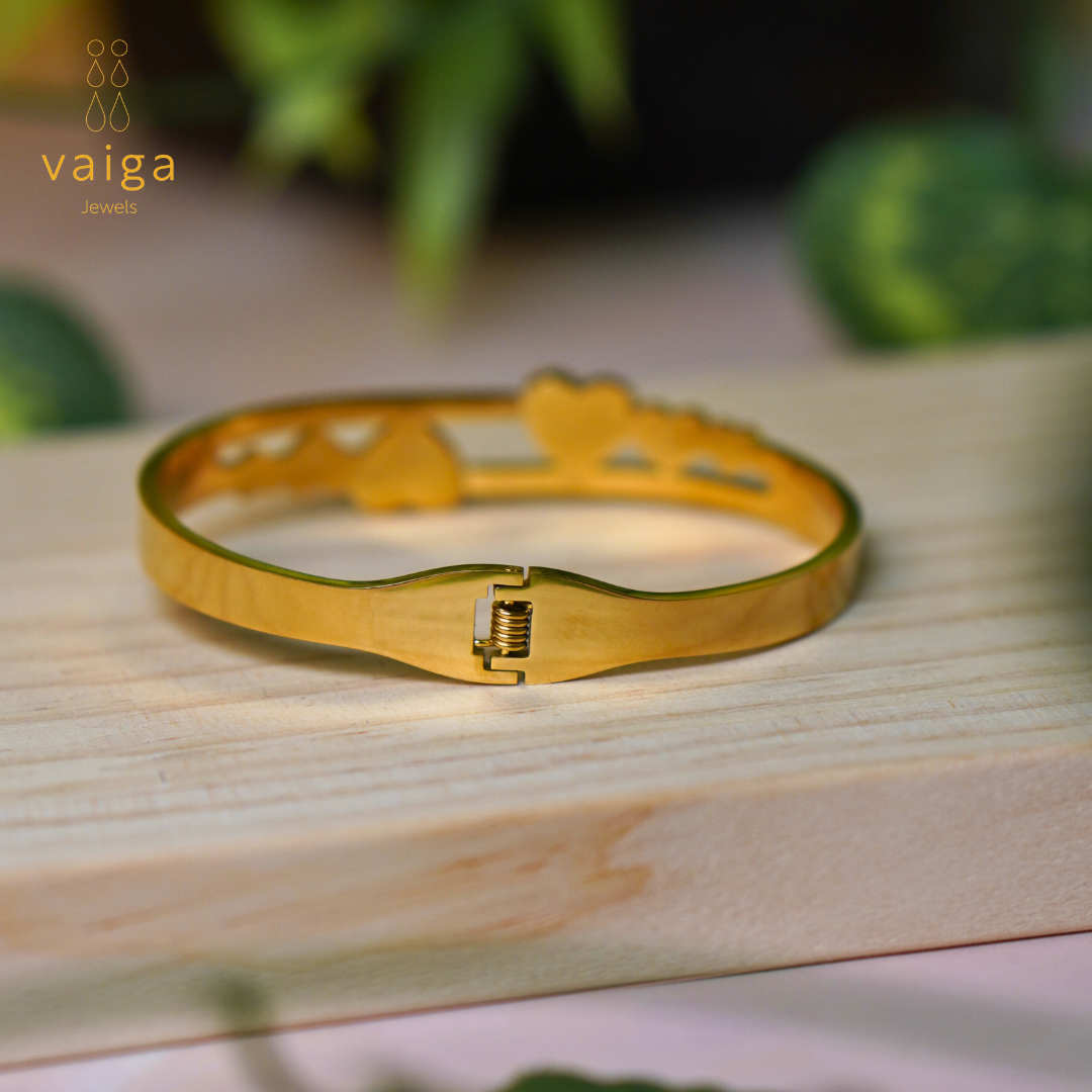 A gold-toned bracelet displayed on a wooden surface, showcasing its secure clasp mechanism. The bracelet features an intricate openwork design with heart-shaped elements in the background. The "Vaiga Jewels" logo is positioned in the upper left corner, complementing the elegant and sophisticated aesthetic of the jewelry piece.