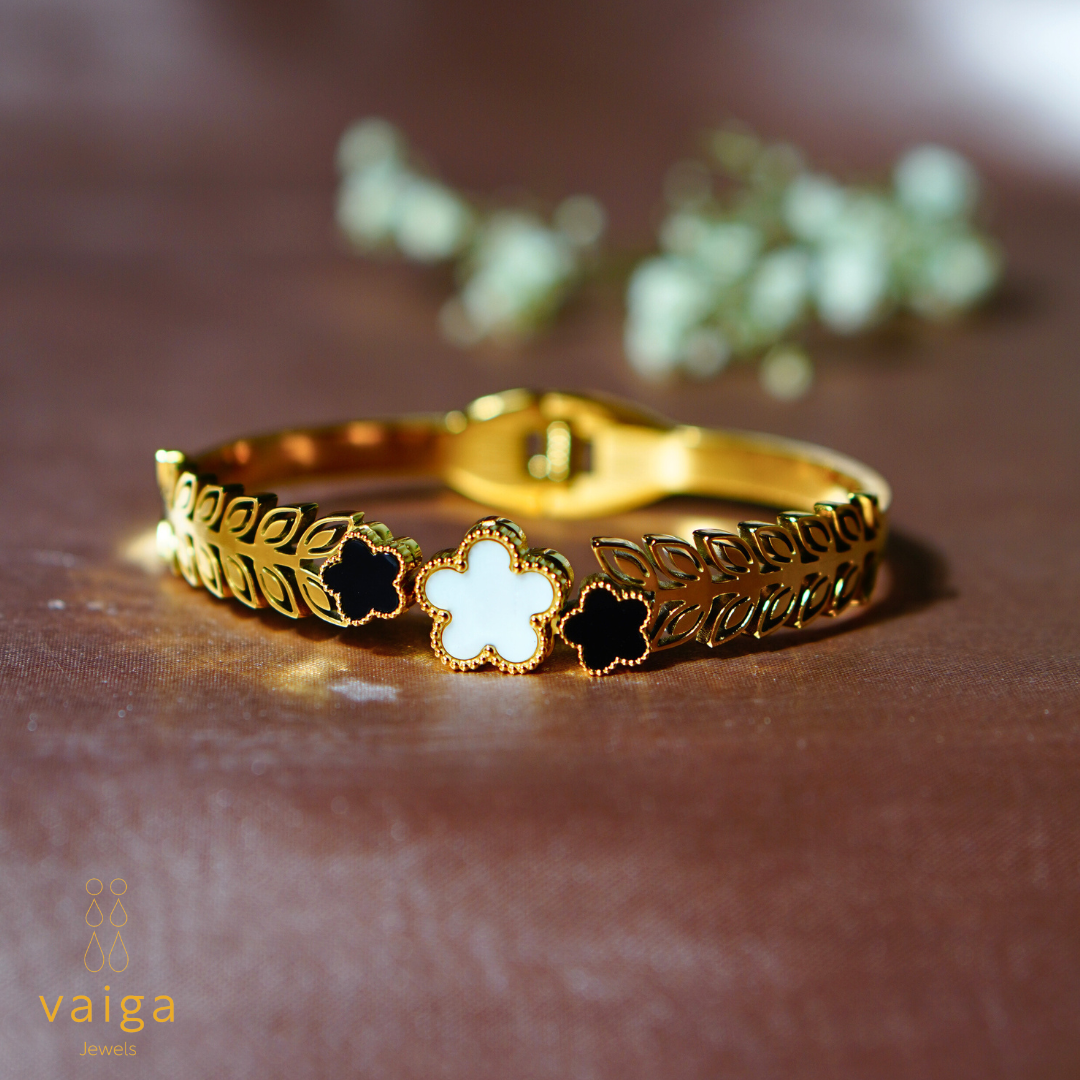 A gold-plated bracelet with an intricate leaf-patterned band and three clover-shaped charms, one in white and two in black, adorned with delicate gold bead detailing. The bracelet features a secure clasp closure. The Vaiga Jewels logo is positioned in the bottom left corner, and the background is a warm brown fabric with softly blurred white flowers for an elegant, luxurious aesthetic.
