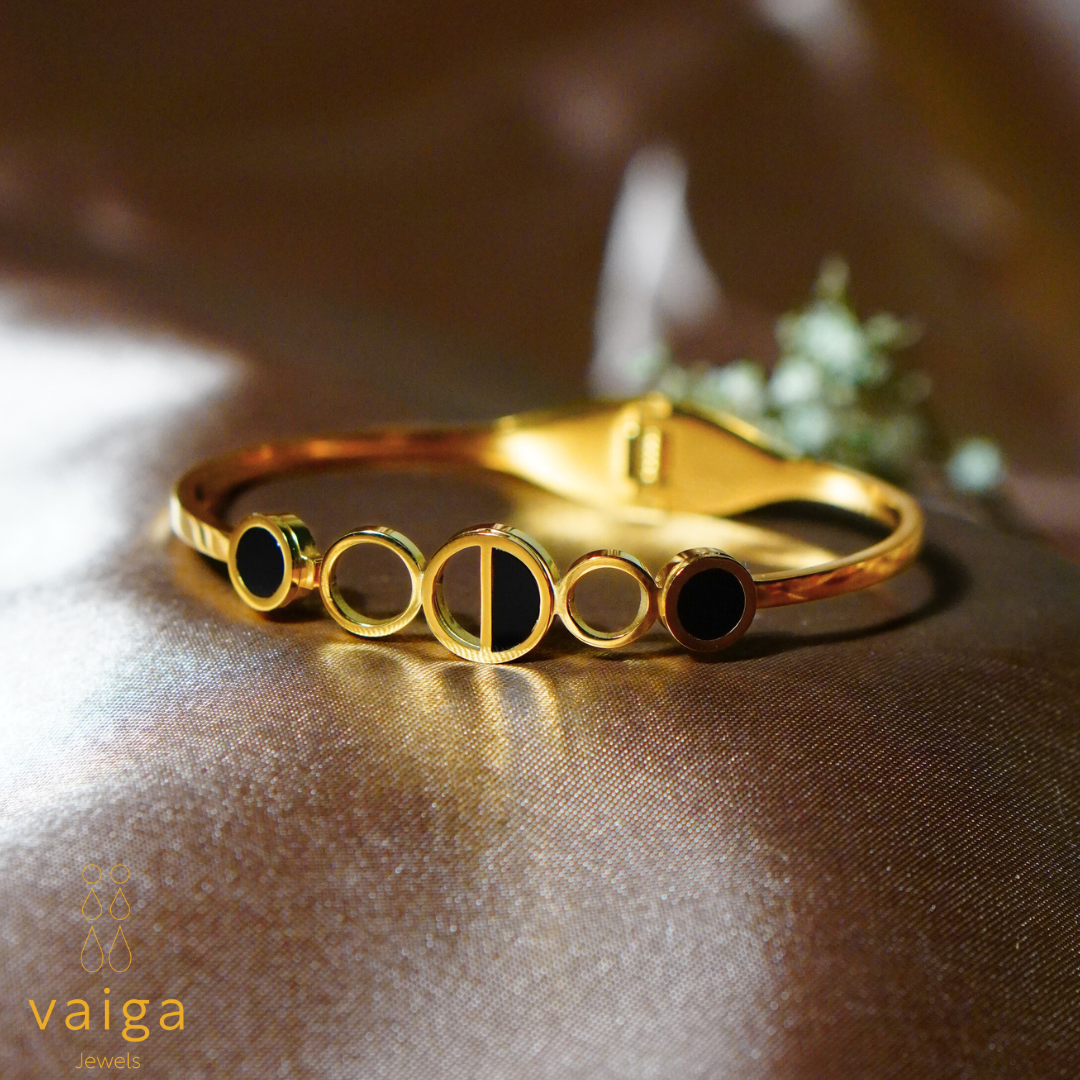 A gold-plated stainless steel bracelet with a modern geometric circle design, featuring black enamel accents. The sleek and minimalist style adds a touch of sophistication. Displayed on a satin background with Vaiga Jewels branding in the corner.