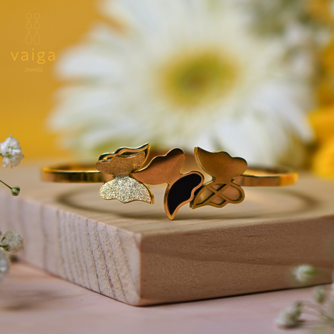  A gold-toned bracelet with intricate butterfly and abstract floral designs, featuring a mix of textured, polished, and black enamel finishes. The bracelet is displayed on a wooden surface with a soft-focus floral background, creating an elegant and premium aesthetic. The "Vaiga Jewels" logo is subtly placed in the upper left corner.