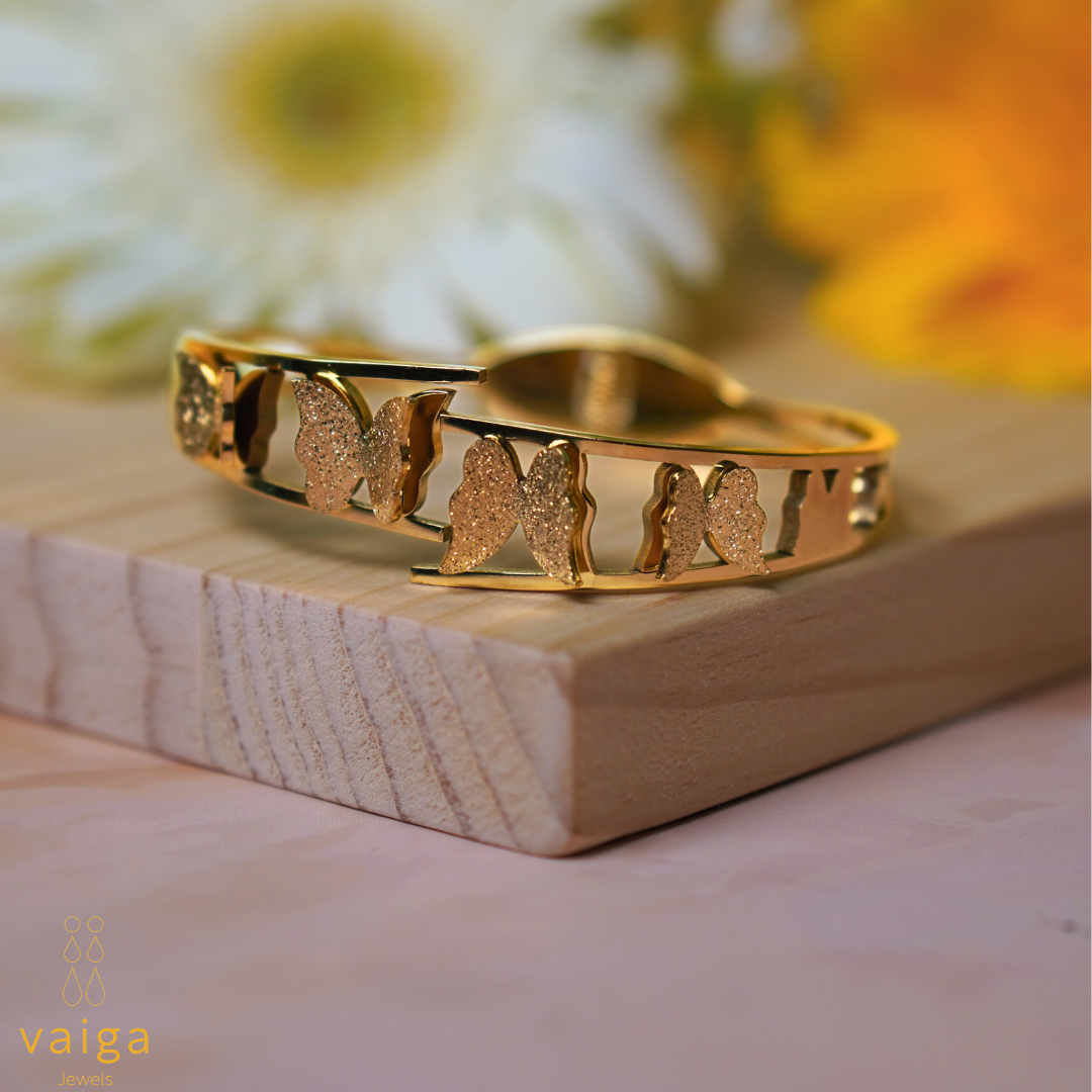  A gold bracelet featuring an intricate butterfly design with textured and polished finishes, displayed on a wooden surface. The background includes soft-focus white and yellow flowers, adding a delicate and elegant touch. The "Vaiga Jewels" logo is visible in the bottom left corner.