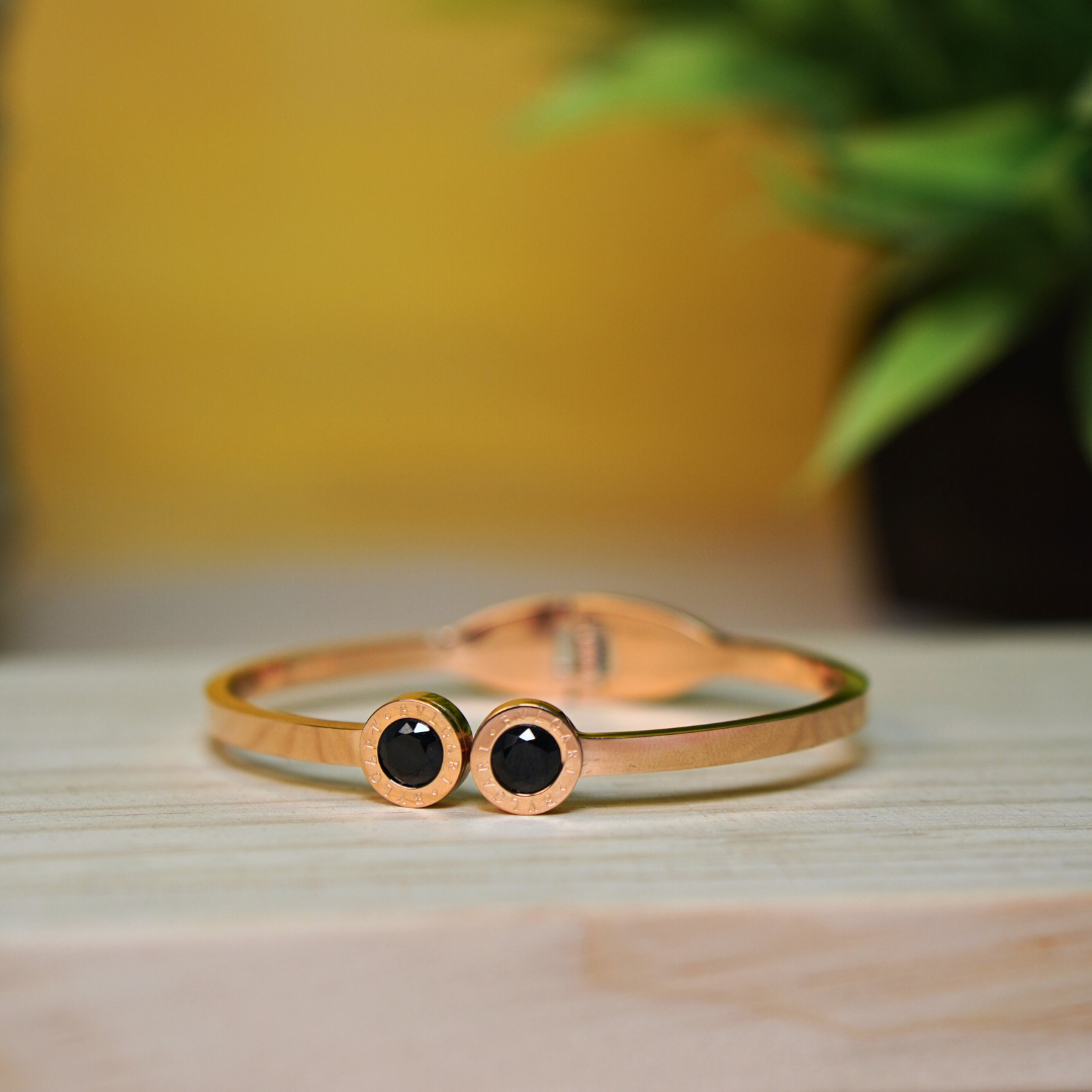 A sleek rose gold bracelet featuring two round black gemstone embellishments, set against a minimalistic wooden surface with a blurred green plant in the background. The bracelet has a modern and elegant design, making it a versatile accessory for various occasions.