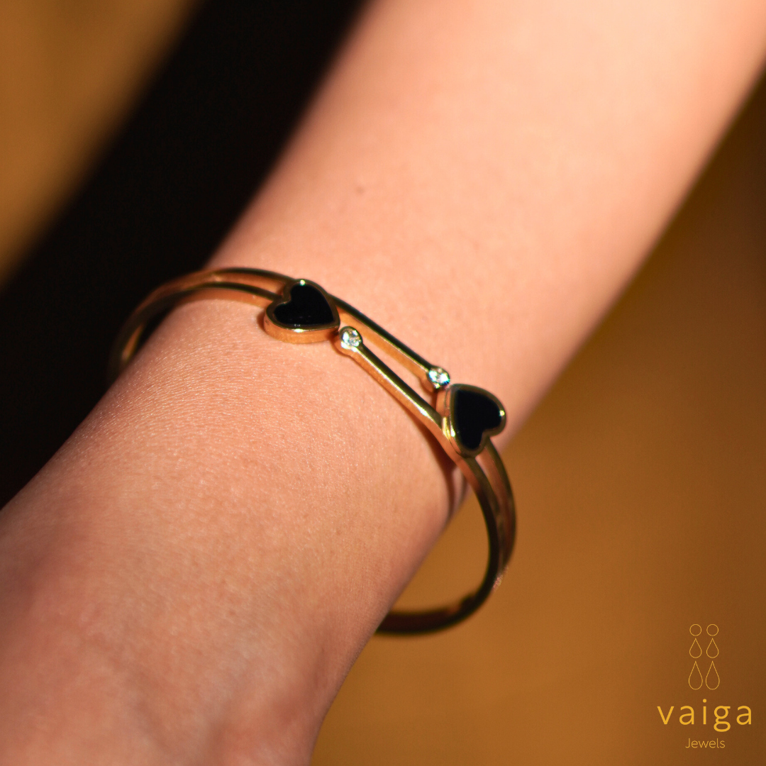 A gold-toned bracelet with black heart-shaped elements and small clear gemstones is showcased on a wrist. The double-band design adds elegance and sophistication. The warm lighting enhances the rich gold hue, complementing the jewelry. The "Vaiga Jewels" logo is visible in the bottom right corner, reinforcing the brand identity.