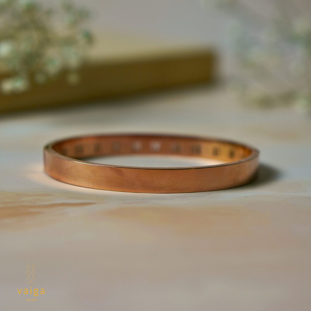 A minimalist rose gold bangle with a smooth, polished exterior and subtle engravings on the inner side, set against a soft, elegant background. The sleek design makes it a versatile and timeless jewelry piece.