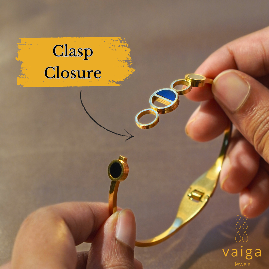 A close-up of hands holding a gold-plated stainless steel bracelet, showing its secure clasp closure mechanism. The bracelet features a modern geometric circle design with black and blue enamel accents. A yellow label with "Clasp Closure" and an arrow highlights the fastening system. The Vaiga Jewels logo is present in the corner.