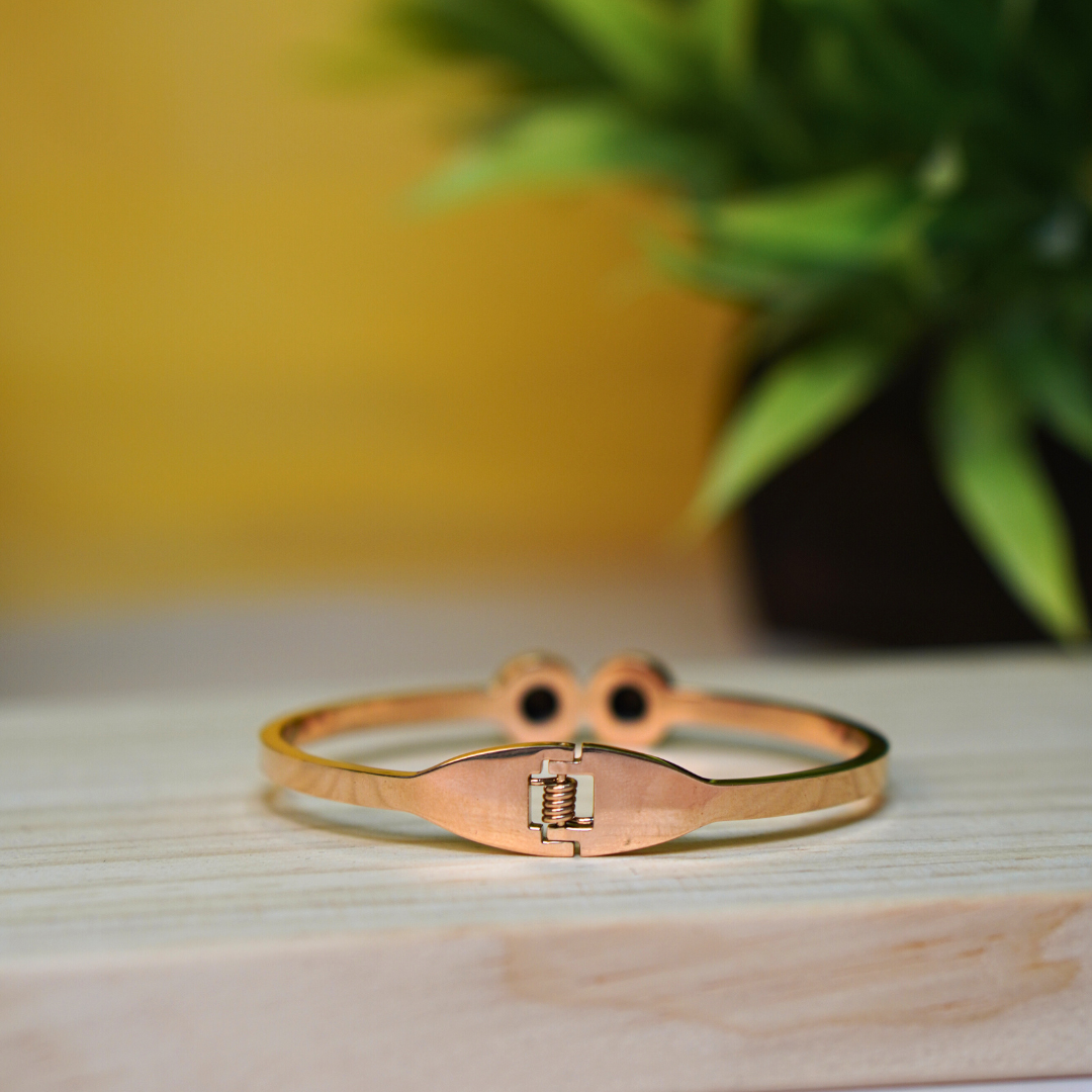  A close-up view of the backside of a rose gold bracelet, highlighting its secure clasp mechanism. The bracelet rests on a light wooden surface, with a blurred green plant in the background, adding a natural aesthetic to the image.