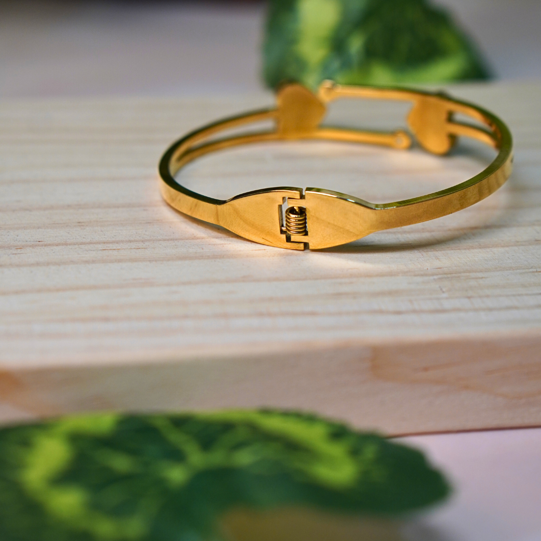 A gold-toned bracelet with a hinge and clasp mechanism, displayed on a light wooden surface. The bracelet has a sleek, polished finish with a secure locking system. Green foliage in the background adds a natural and elegant touch to the image.