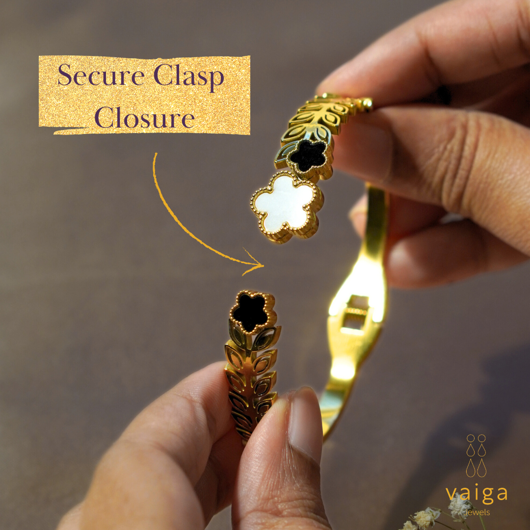 A close-up image of a gold-plated bracelet with a secure clasp closure, being held by two hands. The bracelet features a leaf-patterned band with three clover-shaped charms—one in white and two in black—adorned with delicate gold bead detailing. A golden arrow points towards the clasp, emphasizing its secure mechanism. The phrase "Secure Clasp Closure" is written in an elegant font on a glitter-textured gold banner. The Vaiga Jewels logo is positioned in the bottom right corner. The background is a soft, ne