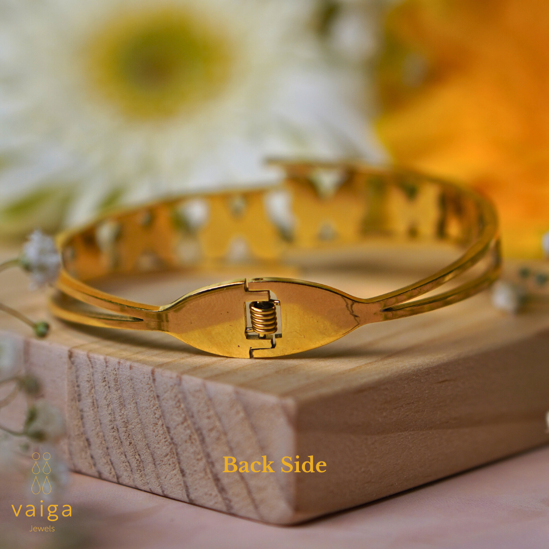 The back side of a gold butterfly bracelet featuring a secure spring clasp mechanism. The bracelet is displayed on a wooden surface with delicate white flowers nearby. A soft-focus background with white and yellow flowers adds a warm, elegant touch. The "Vaiga Jewels" logo and "Back Side" text are visible in the image.