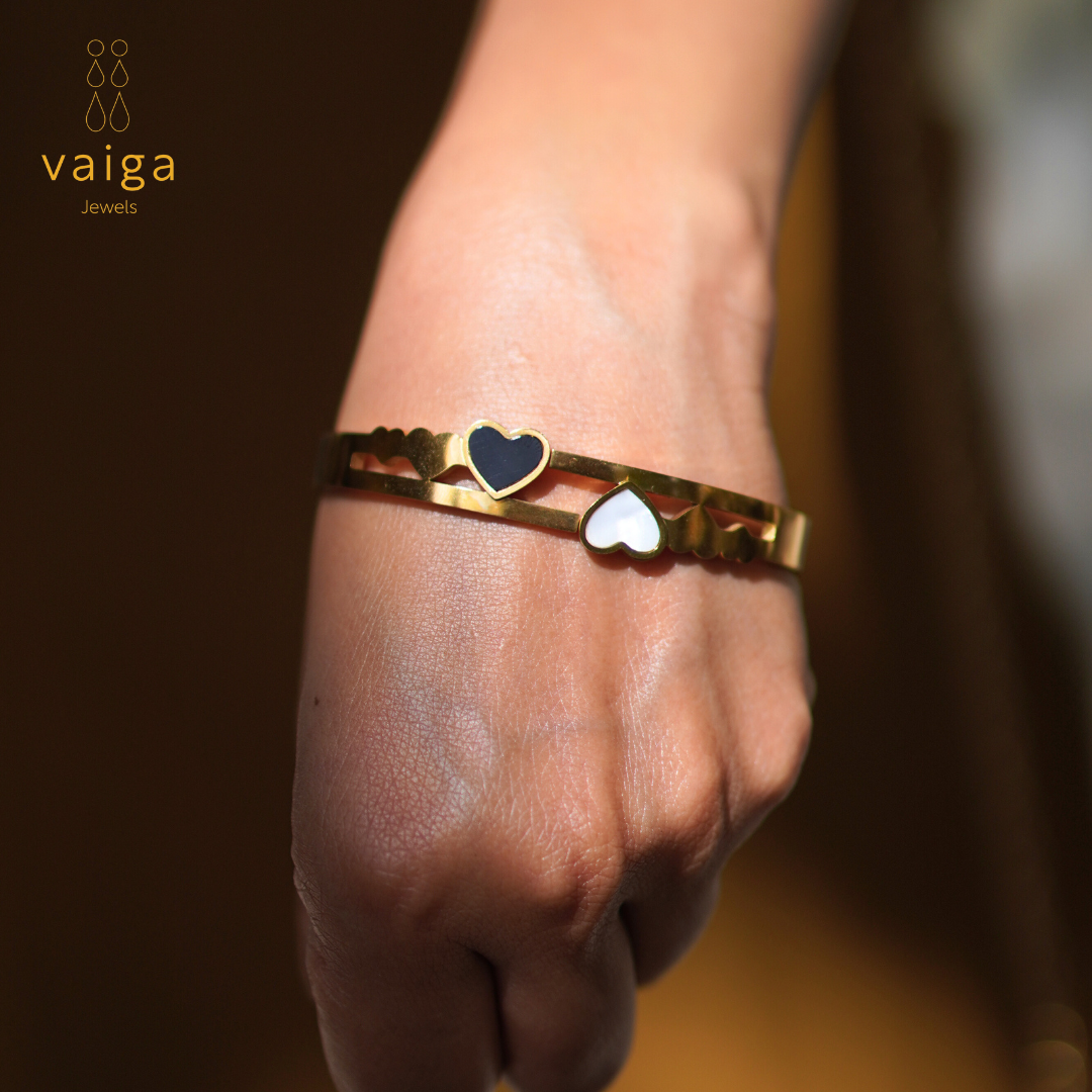  A stylish gold-toned bracelet with black and white heart-shaped accents elegantly worn on a wrist. The openwork design features delicate heart motifs, creating a chic and modern look. The "Vaiga Jewels" logo is visible in the top left corner, enhancing the premium feel of the jewelry piece.
