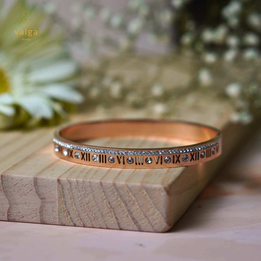A rose gold bangle featuring Roman numerals and sparkling gemstones, elegantly placed on a wooden surface. The bracelet is accented with a row of small crystals along the top, adding a touch of sophistication and luxury. Delicate floral elements in the background enhance the soft, elegant aesthetic.