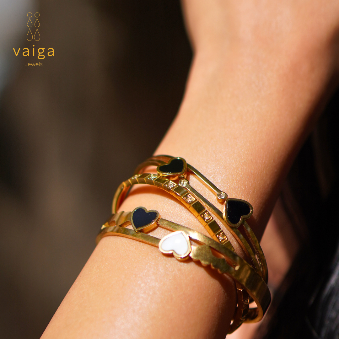A close-up of a wrist adorned with three elegant gold-toned bracelets from Vaiga Jewels. Each bracelet features intricate heart-shaped black and white enamel accents, sparkling stone embellishments, and unique design elements that enhance their charm. The warm sunlight highlights the luxurious finish, creating a radiant and sophisticated look. The "Vaiga Jewels" logo is visible in the top left corner.