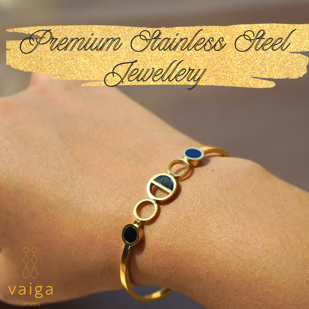 A gold-plated stainless steel bracelet with a modern geometric circle design, featuring black and blue enamel details. Worn on a wrist, showcasing its sleek and stylish fit. The image includes the text "Premium Stainless Steel Jewellery" on a gold glitter banner, along with the Vaiga Jewels logo in the corner.