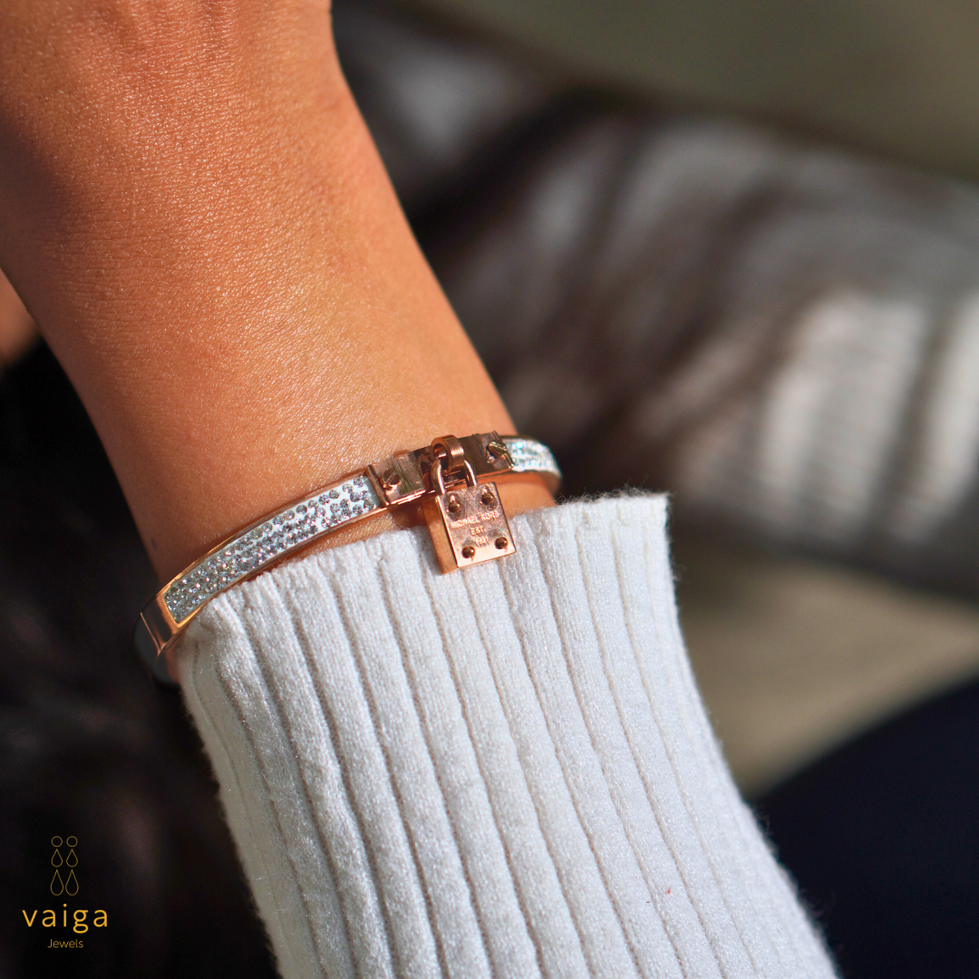 A stylish rose gold bracelet with shimmering crystal embellishments and a lock charm, elegantly worn on a wrist. The close-up shot captures the intricate details of the design, highlighting its luxurious appeal. The soft knit sleeve and blurred background add a cozy, sophisticated touch. The "Vaiga Jewels" logo is subtly placed in the lower left corner.