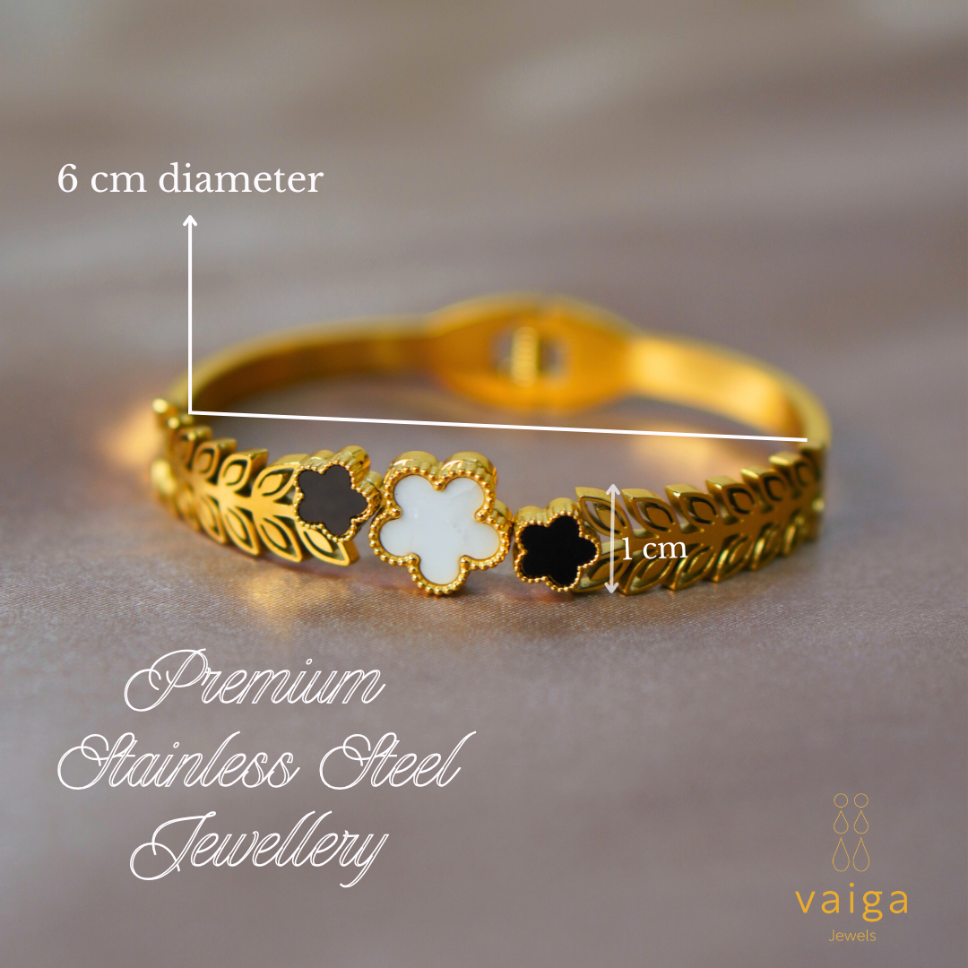 A gold-plated bracelet with a leaf-patterned band and three clover-shaped charms—one in white and two in black—accented with intricate gold bead detailing. The bracelet has a 6 cm diameter and a 1 cm charm height, as indicated by labeled measurements. "Premium Stainless Steel Jewellery" is elegantly written in cursive font, and the Vaiga Jewels logo is positioned in the bottom right corner. The background features a soft, neutral fabric for a refined and luxurious aesthetic