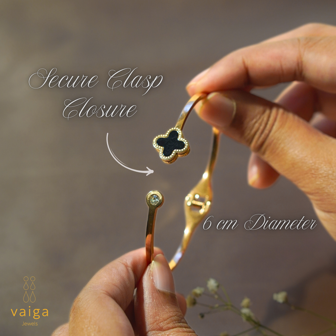 A close-up of a rose gold-plated bracelet being held in both hands, showcasing its secure clasp closure. The bracelet features a black clover charm with gold bead detailing and a sparkling crystal accent at the clasp. The image includes elegant text highlighting the "Secure Clasp Closure" and "6 cm Diameter." The Vaiga Jewels logo is placed in the bottom left corner, and the background has a soft, neutral tone with delicate white flowers for an elegant touch.