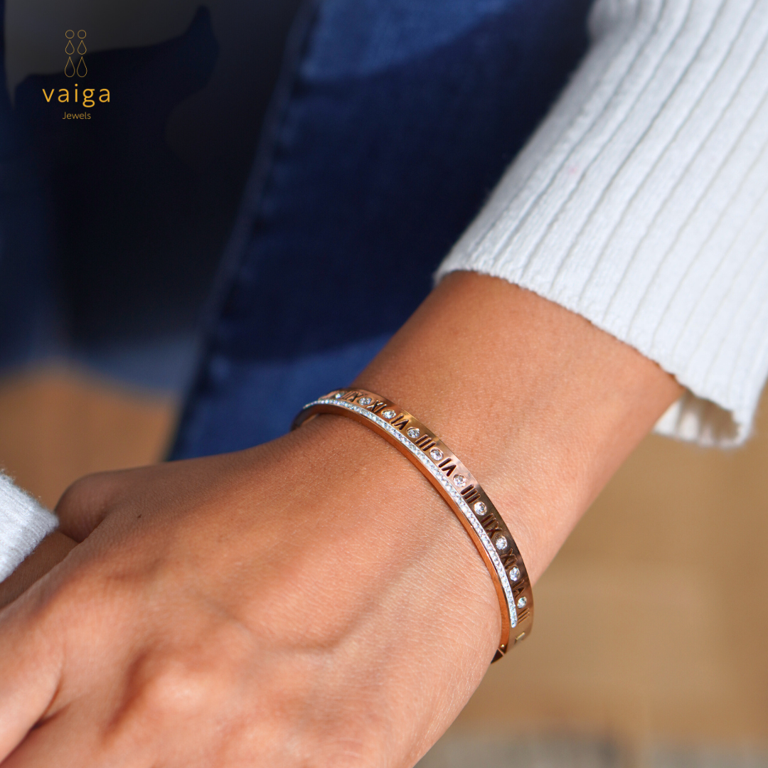 A stylish rose gold bangle with Roman numeral engravings and embedded gemstones, elegantly worn on a wrist. The bracelet is complemented by a row of sparkling crystals, adding a touch of sophistication. The wearer is dressed in a white ribbed sweater and blue denim, creating a chic and modern look. The logo "Vaiga Jewels" is subtly placed in the upper left corner.