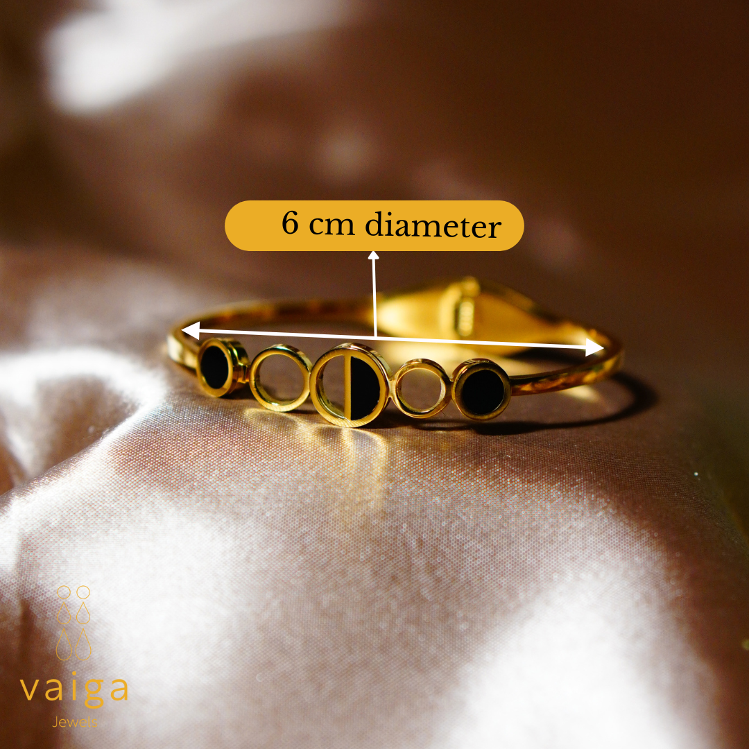 A gold-plated stainless steel bracelet with a modern geometric design, featuring black enamel accents. The bracelet is displayed on a luxurious satin background, with an overlay label indicating its 6 cm diameter. The Vaiga Jewels logo is positioned in the bottom left corner.