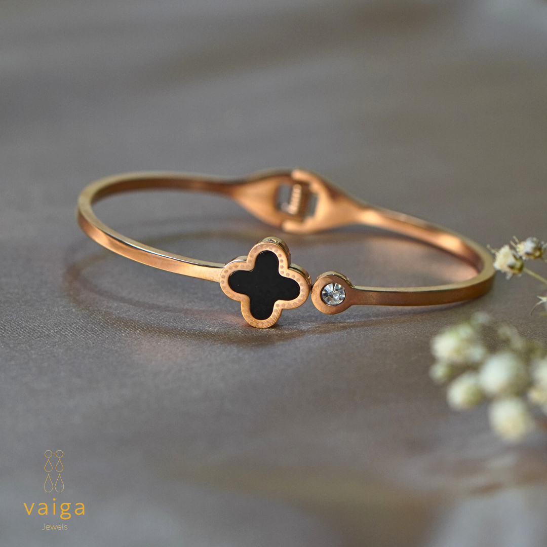 A rose gold-plated stainless steel bracelet featuring a black clover-shaped centerpiece and a single sparkling crystal accent. The bracelet is elegantly displayed on a soft, neutral fabric background with delicate floral elements, complementing its sophisticated design. The Vaiga Jewels logo is positioned in the bottom left corner