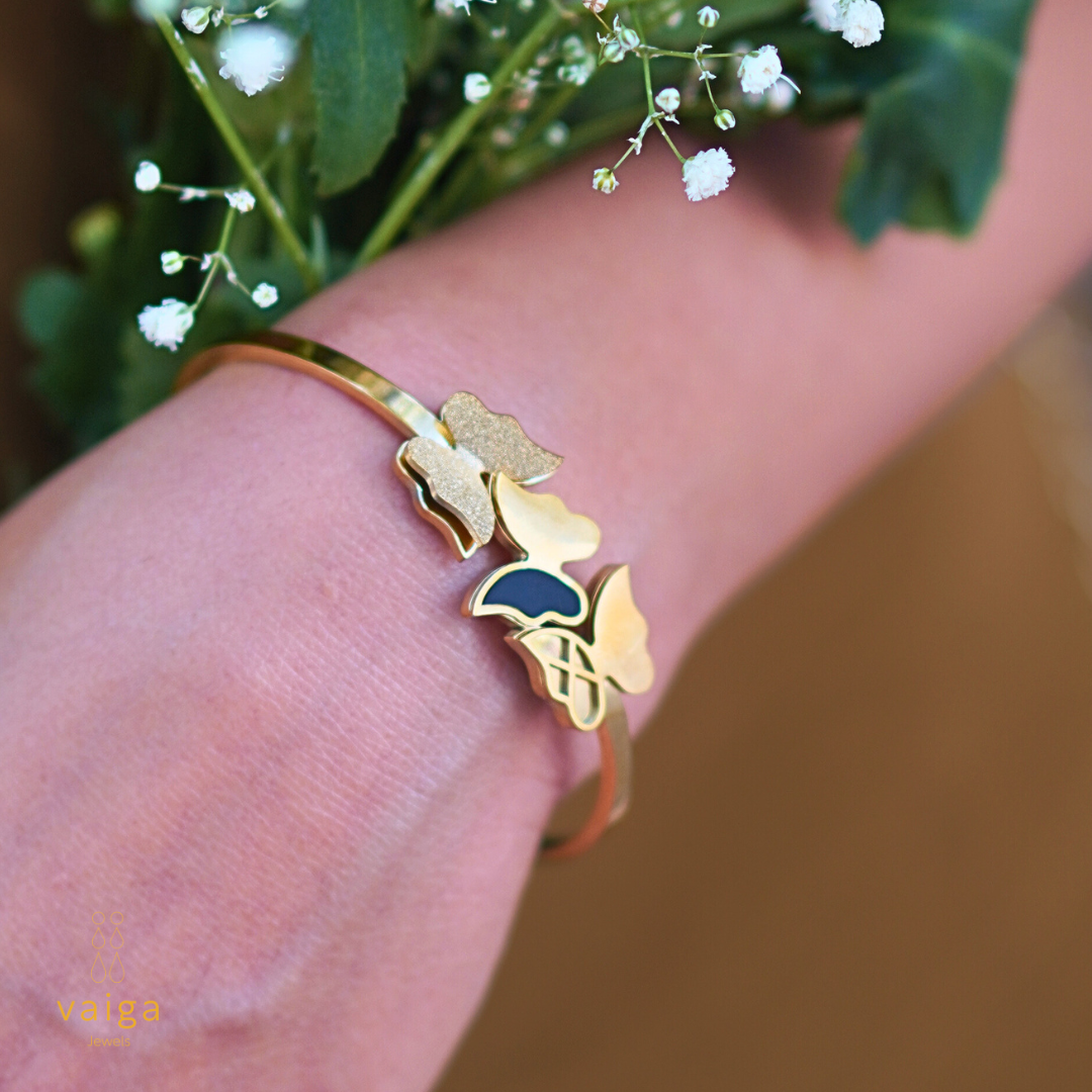 A gold-toned bracelet with a butterfly and floral design, featuring textured, glossy, and black enamel details, elegantly worn on a wrist. The wearer holds a bouquet of delicate white flowers, adding a soft and natural touch to the composition. The "Vaiga Jewels" logo is subtly placed in the lower left corner.