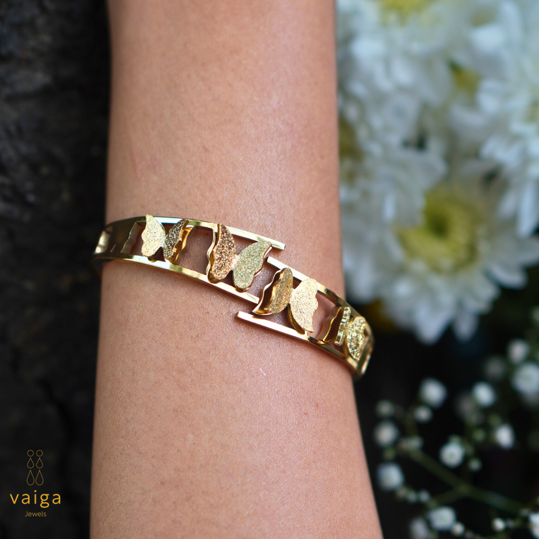  A gold butterfly bracelet elegantly worn on a wrist, showcasing its intricate cut-out design with textured and polished butterfly motifs. The background features delicate white flowers, enhancing the bracelet's sophisticated and feminine appeal. The "Vaiga Jewels" logo is visible in the bottom left corner.