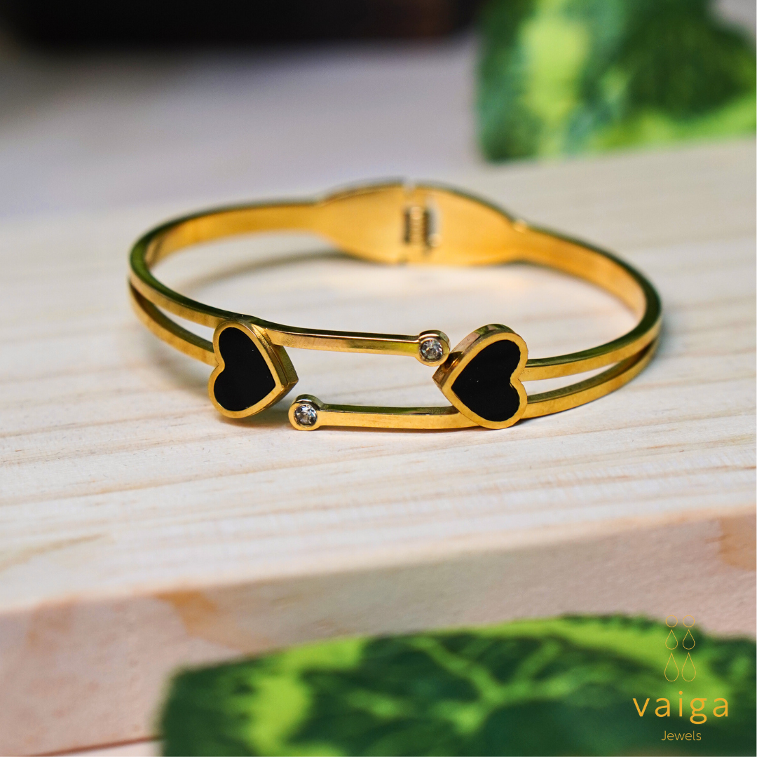 A gold-toned bracelet with a double-band design featuring two black heart-shaped elements at each end, accented by small clear gemstones. The bracelet is displayed on a light wooden surface with green foliage in the background, creating a natural and elegant aesthetic.