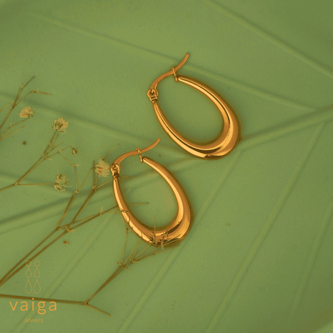 Gold-toned stainless steel tear drop-shaped  earrings by Vaiga Jewels, modern luxury women's accessory on green, leaf textured background. Vaiga Jewels logo at the bottom left.