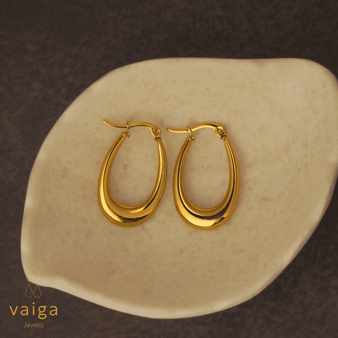 Gold-toned stainless steel tear drop-shaped earrings design by Vaiga Jewels, modern luxury women's accessory placed in a white shell shaped dish . Vaiga Jewels logo at the bottom left.