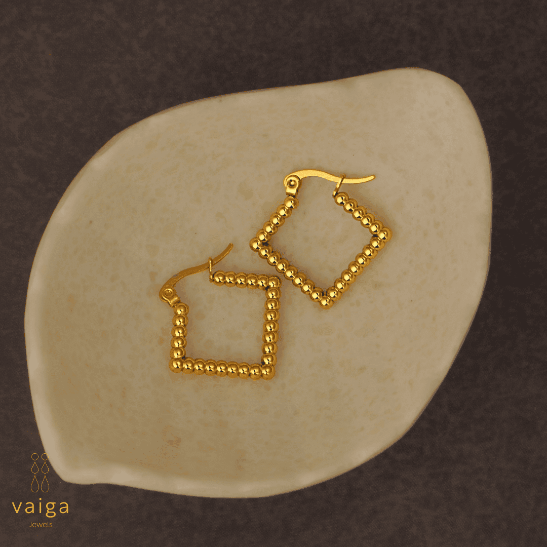 Gold-toned stainless steel  textured square-shaped earrings design by Vaiga Jewels, modern luxury women's accessory placed in a white shell shaped dish . Vaiga Jewels logo at the bottom left.