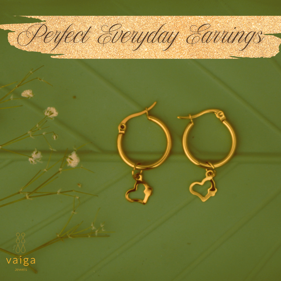Gold-toned stainless steel  hoop earrings with heart shaped charm by Vaiga Jewels, modern luxury women's accessory on green, leaf textured background. Vaiga Jewels logo at the bottom left.