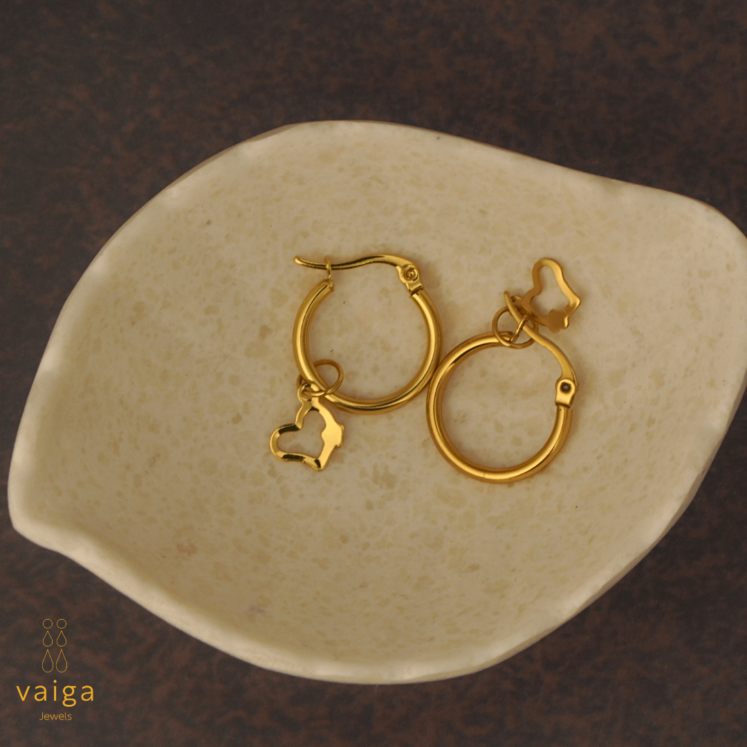 Gold-toned stainless steel hoop earrings with heart shaped charm by Vaiga Jewels, modern luxury women's accessory placed in a white shell shaped dish . Vaiga Jewels logo at the bottom left.